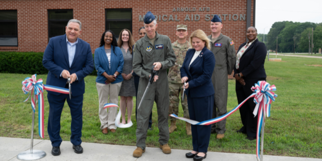 Group of people in suits cutting ribbon in front of brick building.