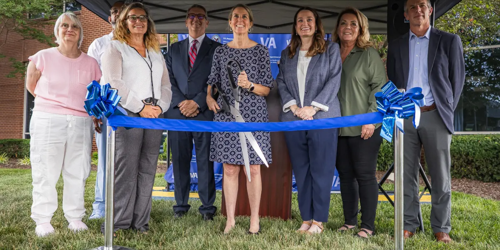 Group of people in formal wear stand behind ribbon at ribbon cutting ceremony.