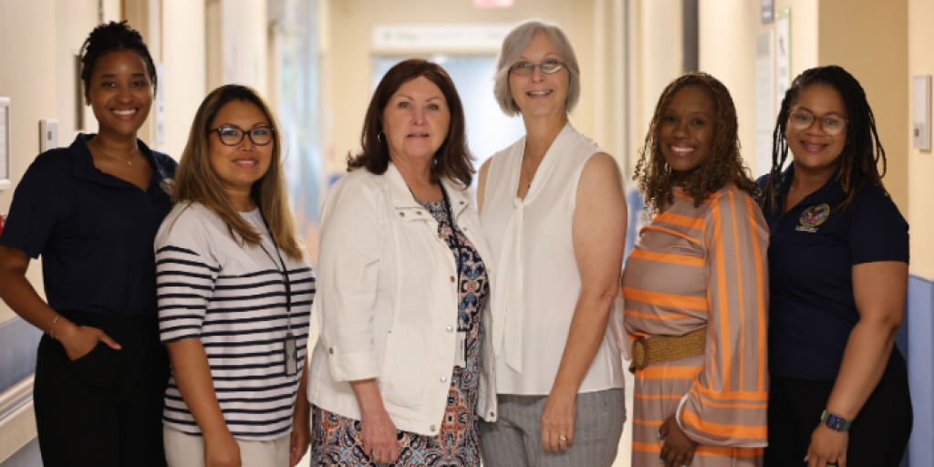 Six women stand in a hospital corridor. Four wear uniforms, two in casual attire.