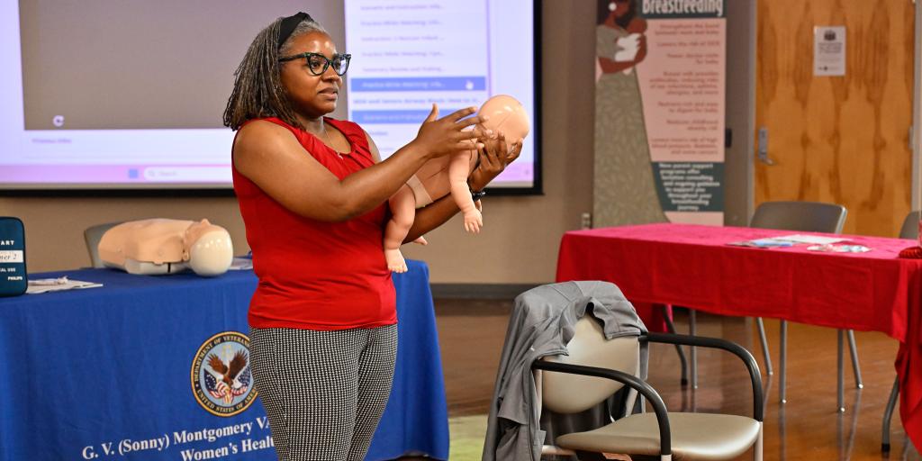 Woman in red shirt holding baby mannequin in training room.