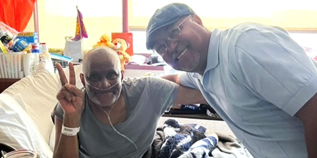 Two men in a hospital room, one making a peace sign.