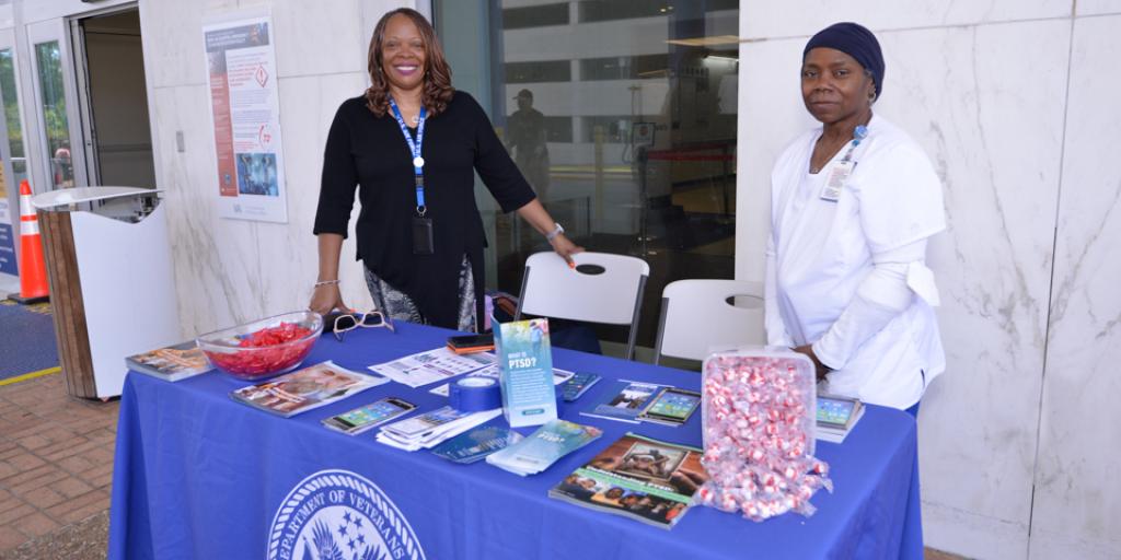 Two women stand behind a blue table with brochures and candy, outside a building.