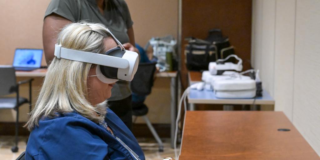 Woman in blue scrubs using VR headset and controller, supervised by another woman.