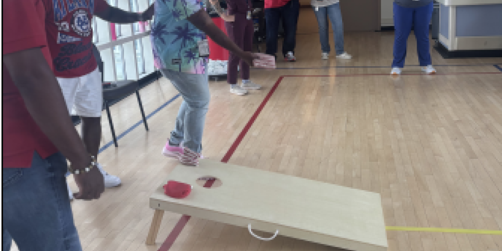 Man in tie-dye shirt throws beanbag onto cornhole board during game in gym.