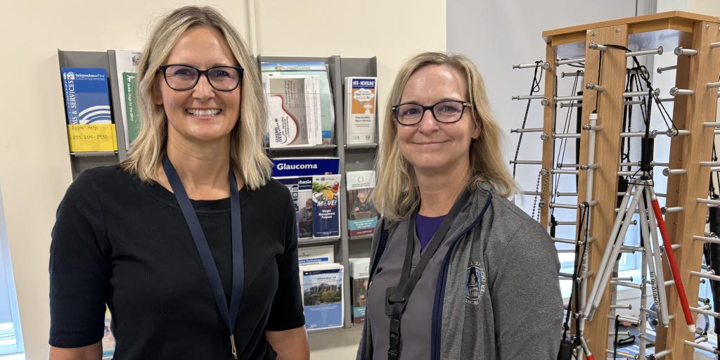 Two women in glasses and staff badges stand in front of a bookshelf and medical equipment.