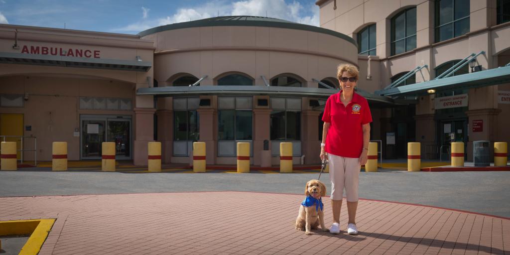 Woman in red shirt and white pants with service dog in front of hospital.