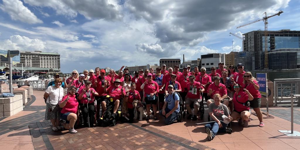 Group of people in wheelchairs and standing, wearing pink shirts, pose on a brick plaza.