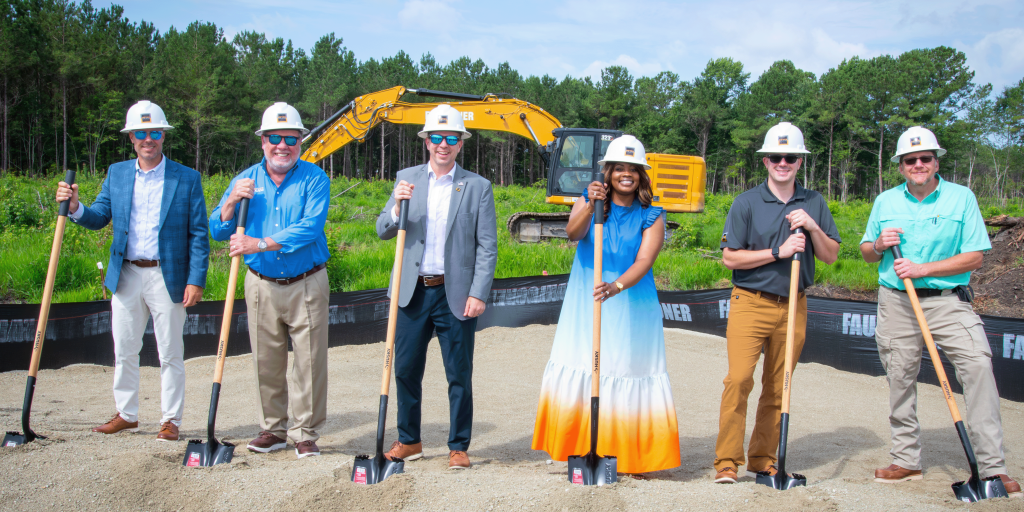 Six people in hard hats hold shovels at a groundbreaking ceremony.