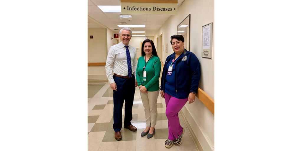 Three people in a hospital hallway: man in tie, woman in green shirt, woman in blue jacket.