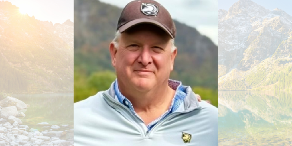 Portrait of a man in a white shirt and brown cap, smiling against a scenic mountain backdrop.