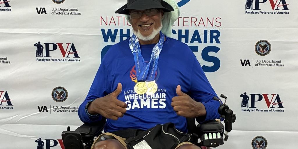 Veteran athlete in wheelchair giving thumbs up with medals on, standing in front of Veterans Wheelchair Games banner.