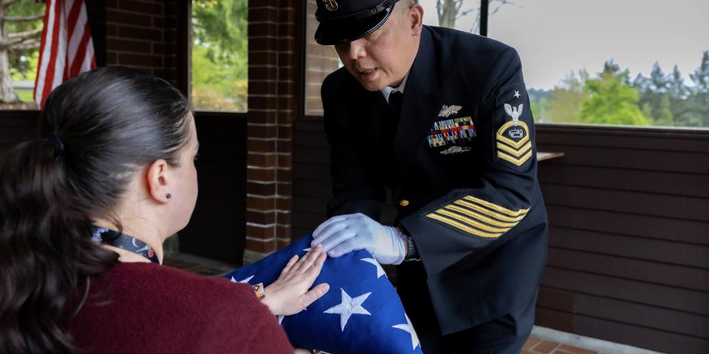 A sailor in uniform presenting a folded flag to a woman.