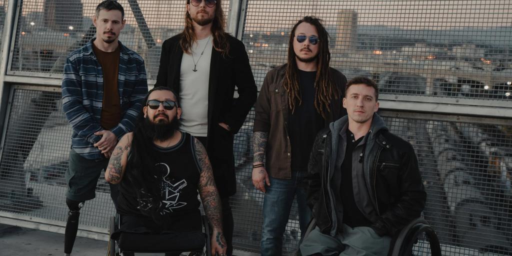 Five men, two in wheelchairs, pose on a bridge with city skyline in background.