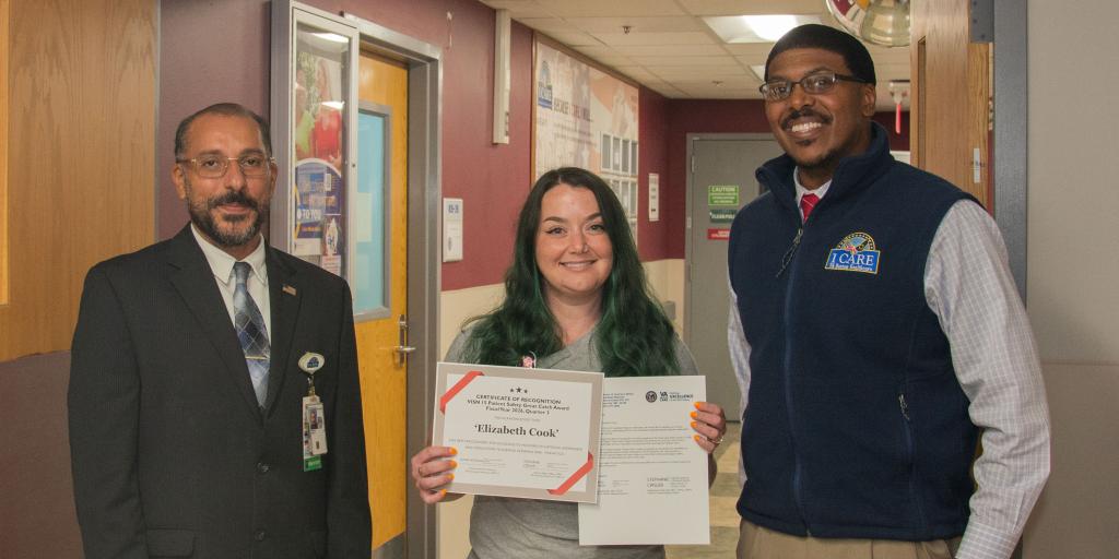 Woman holding a certificate between two men in a school hallway.