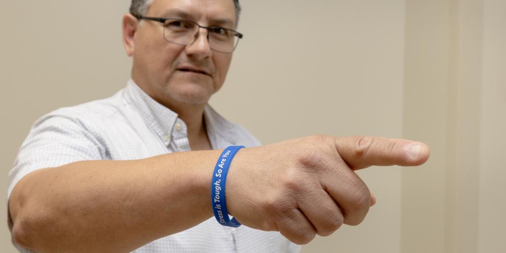 Man in white shirt pointing with blue wristband that says "Stress is tough, so are you"