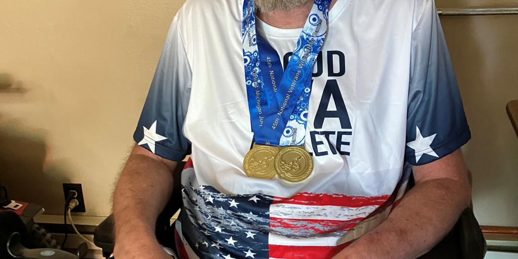 A man with a beard in a wheelchair wearing a USA flag t-shirt and medal.