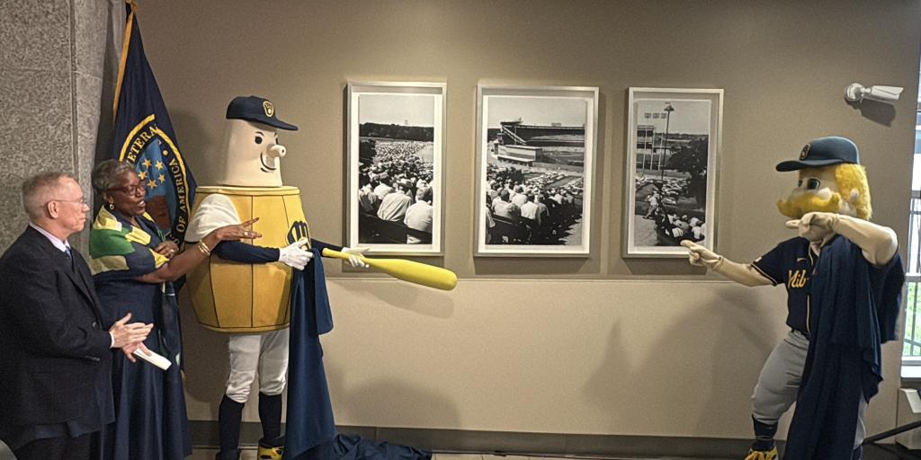 Two mascots in blue robes hold a bat as a man looks on.