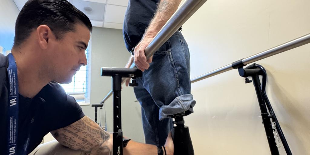 Two men in a hospital room, one helping the other walk with a prosthetic leg.