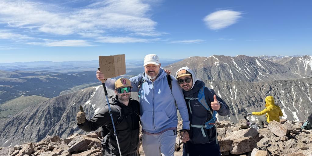 Three hikers on mountain peak, one holding sign, with scenic view.