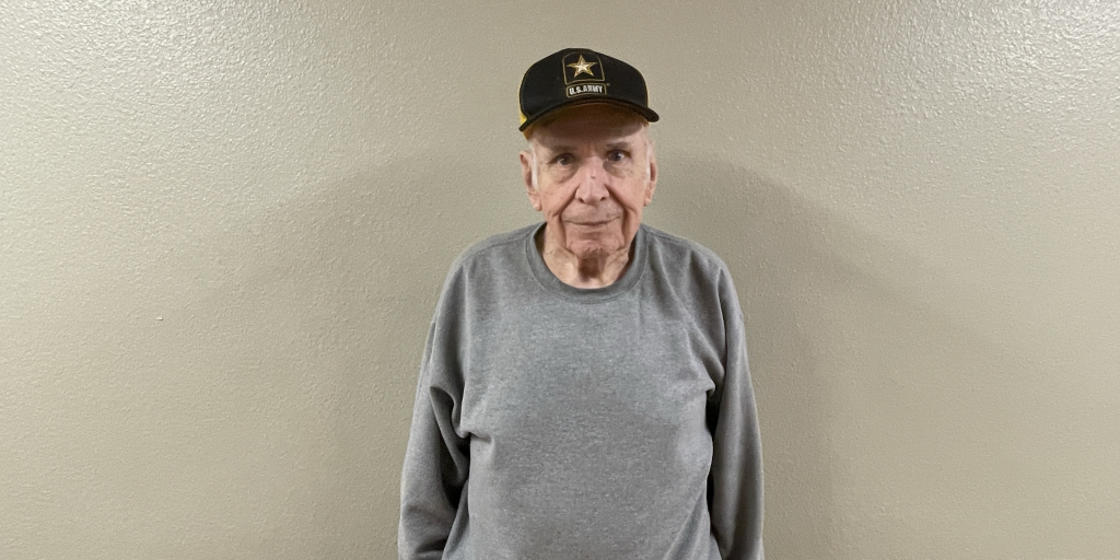 Army Veteran John Wasson, who wrote a song for his VA care team, stands against a light-colored wall wearing an Army cap.