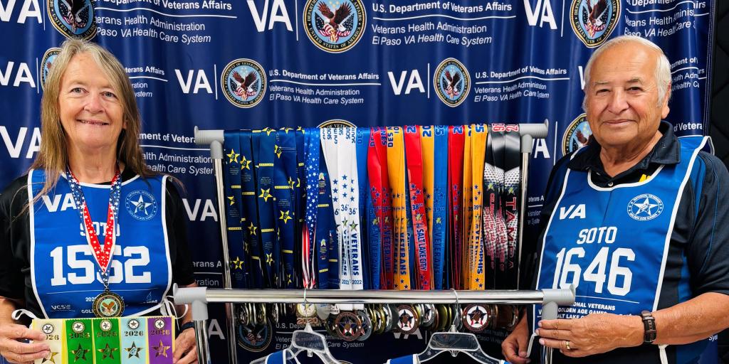 Two smiling people stand next to a rack of medals and ribbons.