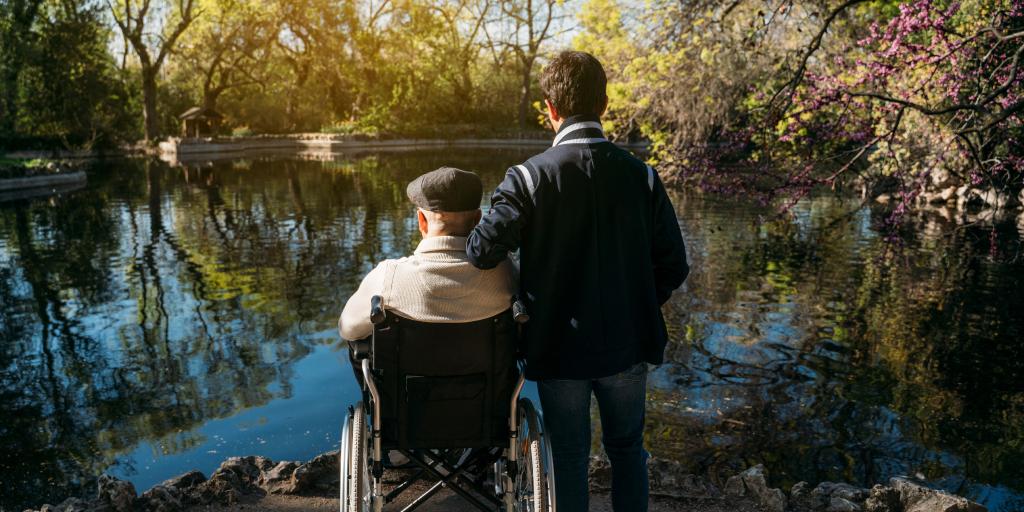 A younger man supports an older man in a wheelchair by a pond.