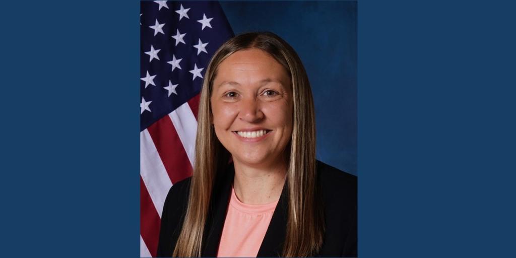 Smiling woman in pink shirt and black jacket in front of US flag.