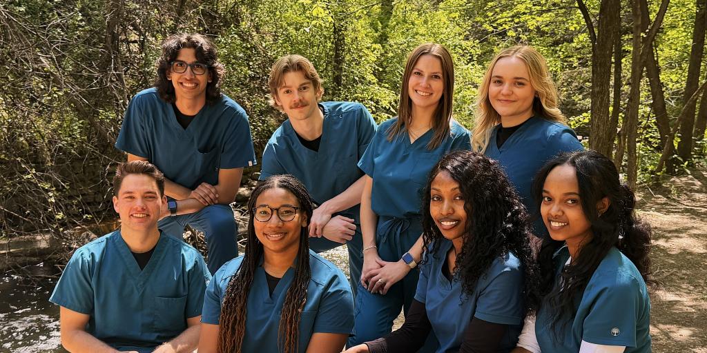 Group of seven dental staff in teal scrubs smiling in forest setting.