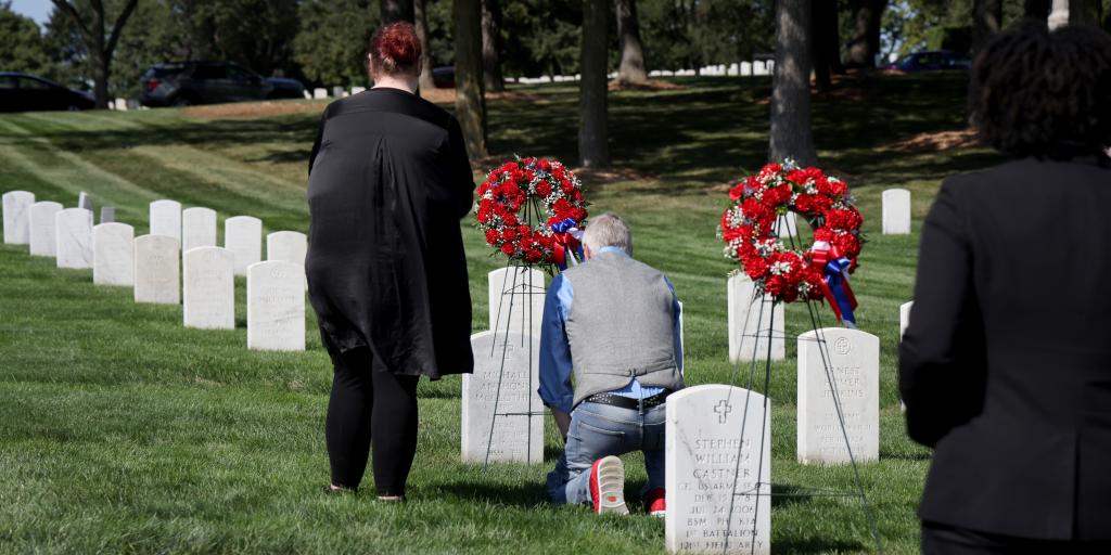 Man kneels at grave with two red wreaths, observed by two others in black.