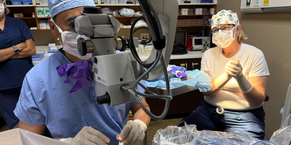 A nurse volunteer sits beside an eye surgeon looking through a surgical microscope during a procedure.