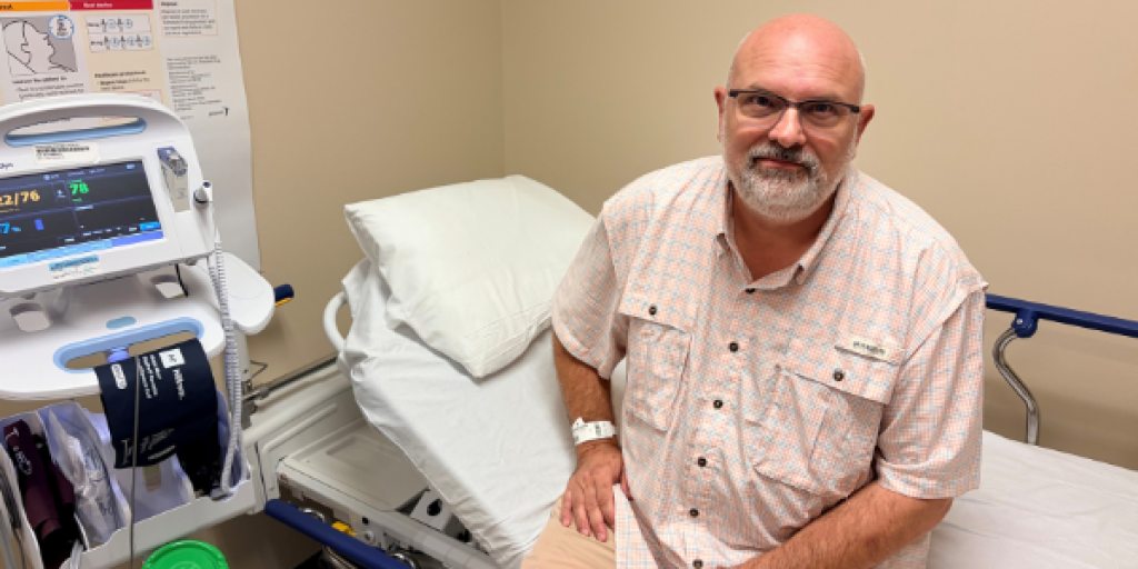 Man with glasses and a beard sitting on a hospital bed next to a monitor.