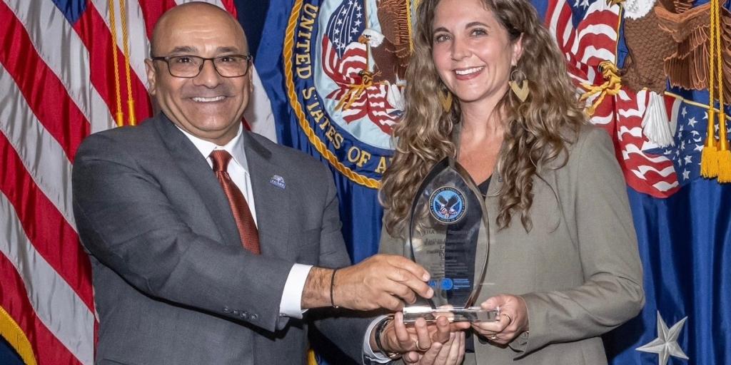 Man presents an award to a smiling woman in front of flags.