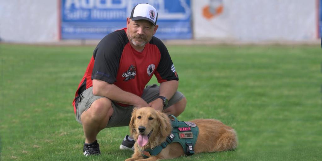 Man kneeling on grass with a service dog beside him.