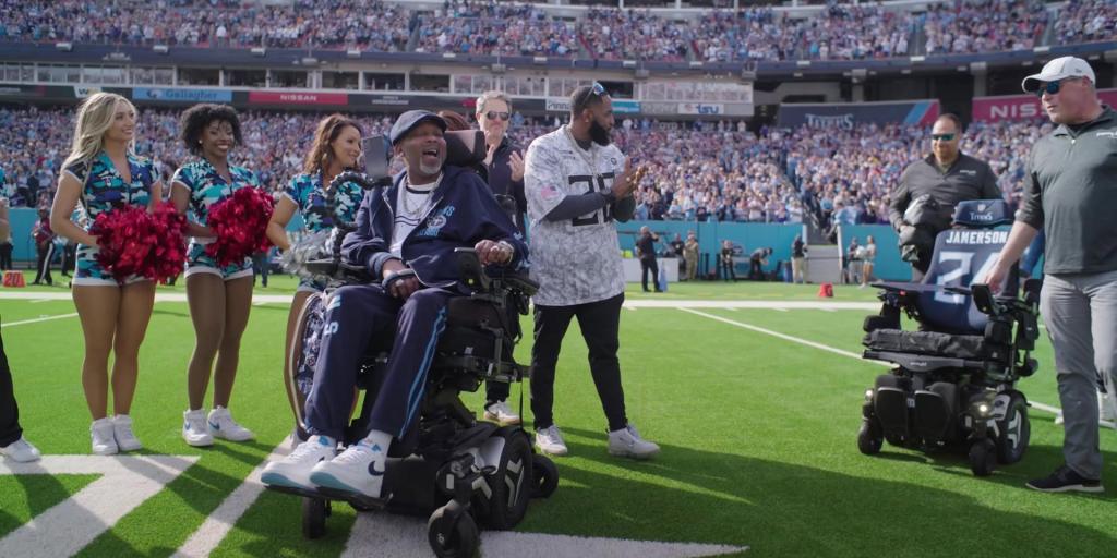 Man in wheelchair on football field with cheerleaders and crowd in stands.