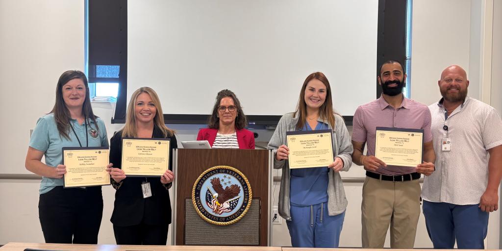 Six people stand behind a podium holding certificates in a classroom.