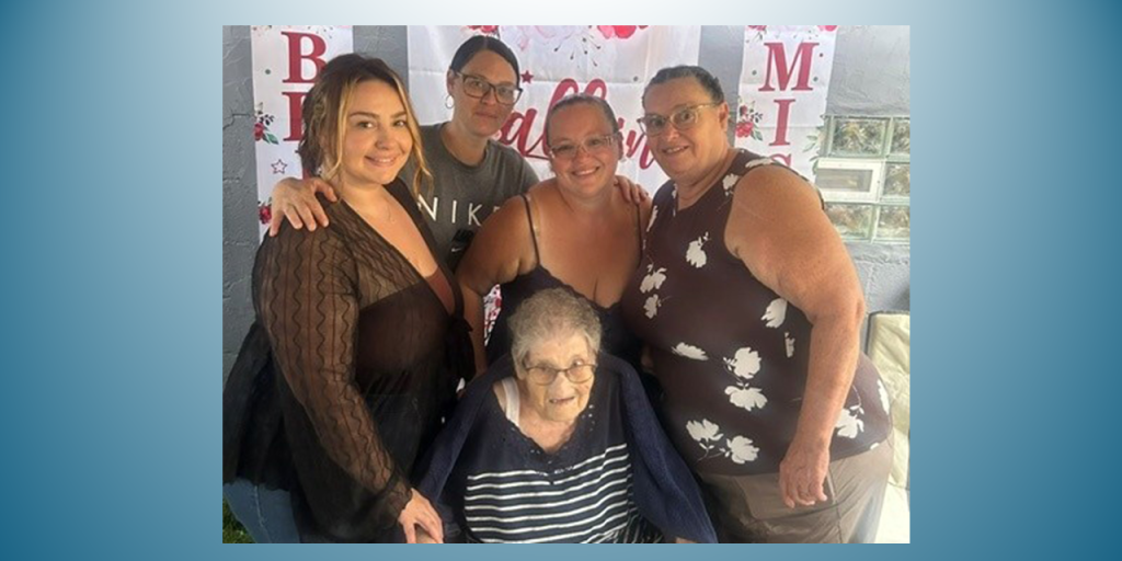Five people stand together in a photo, smiling, with an elderly woman in the center.