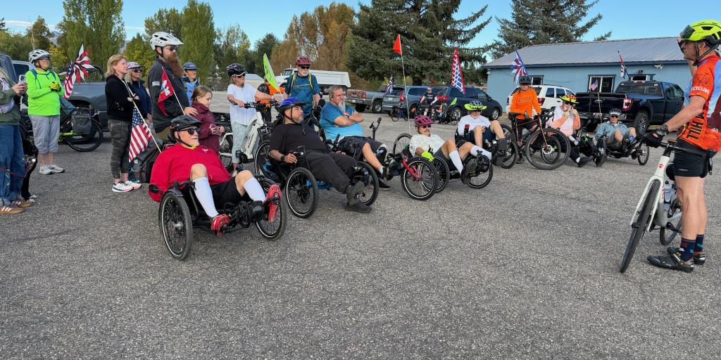 Cyclists in adaptive bikes gather in a parking lot, with spectators and flags.