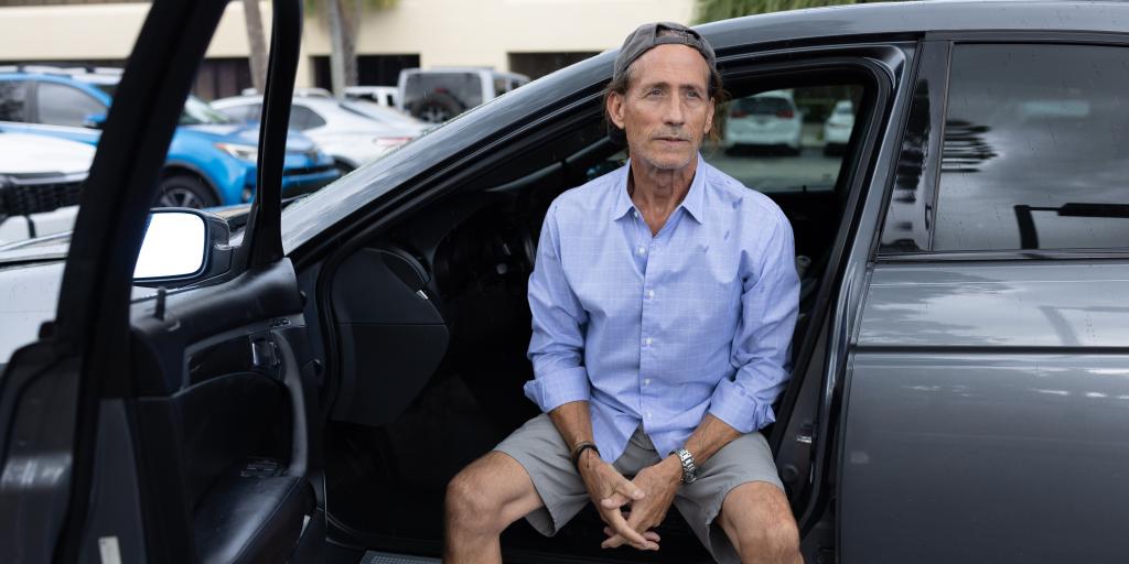 Man in blue shirt and grey shorts sitting in the back seat of his grey car.
