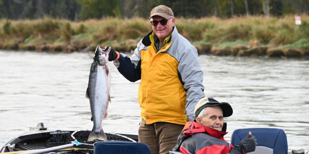 Two men on a boat, one holding a salmon, both smiling.