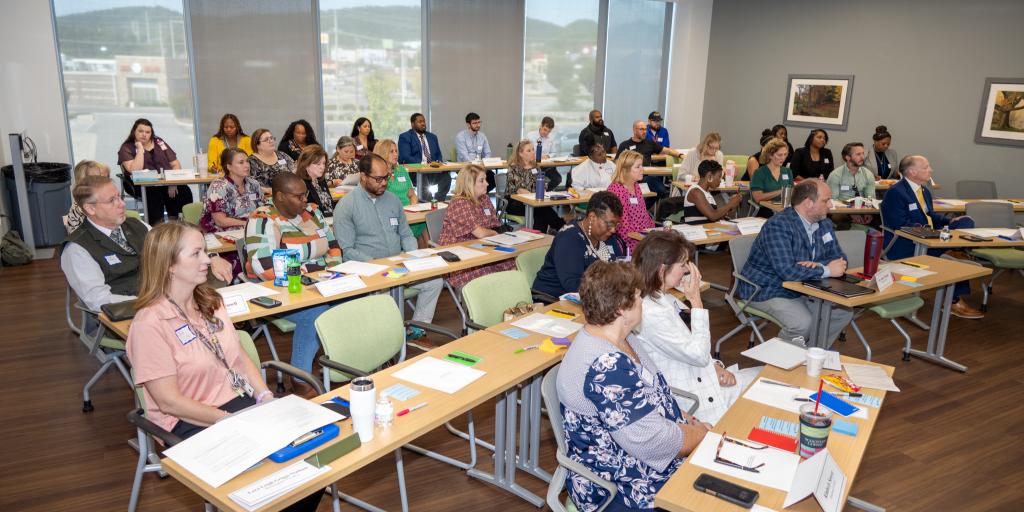 Diverse group of people seated at tables in a conference room with large windows.