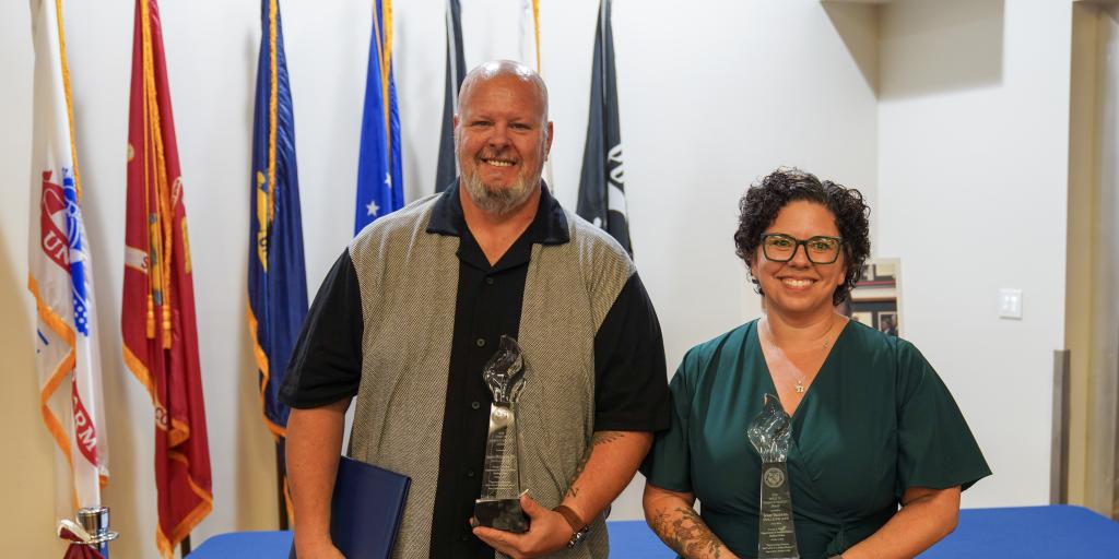Man and woman holding awards in front of flags.