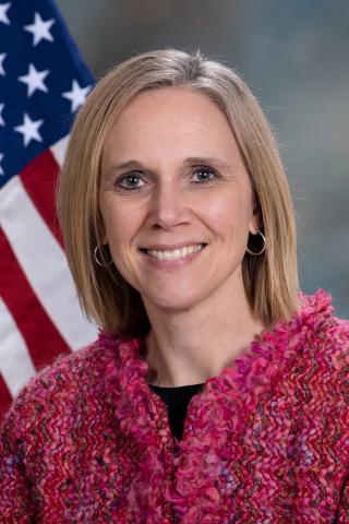 photo of a person with shoulder length, blond hair, gold hoop earrings, wearing a pink boucle jacket in front of a medium gray background and an American flag