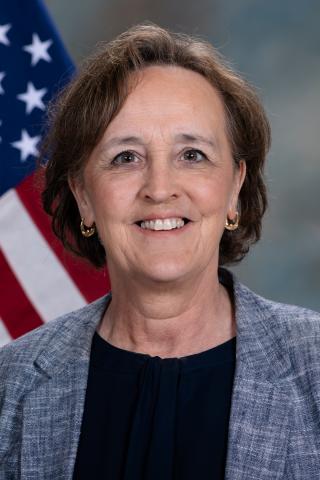 photo of person smiling with chin length brown hair, gold hoop earrings, wearing a light gray jacket over a black shirt, the background is medium gray with an American flag