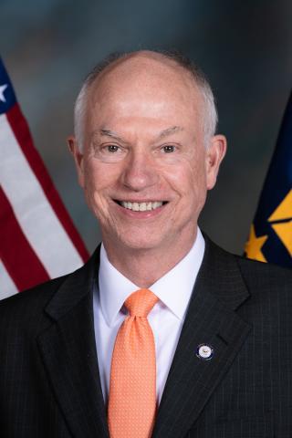 Photo of Patrick J Kelly - headshot of a person with closely cropped gray hair and smiling, they are wearing a dark gray suit with a white shirt and peach colored tie, there are flags behind the person