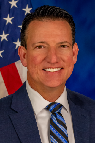 Portrait of Sean N. Turner, Associate Director for the Bay Pines VA Healthcare System. He is smiling directly at the camera and wearing a blue suit with a stripped tie. Directly over his left shoulder is an American Flag.