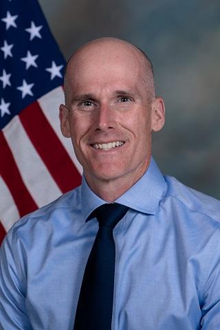 portrait of Michael T. Armstrong, headshot photo of a person with closely cropped hair, smiling at the camera. They are wearing a light blue dress shirt and a navy tie. There is an American flag in the background.