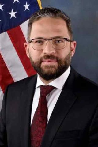 A man dressed in a black suit, white shirt, and red tie, formally posed for an executive-style portrait in front of the American flag
