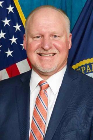 A man in a suit and tie in a formal executive pose in front of the American flag