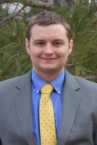 Steve in a grey suit in front of evergreens. 