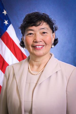 Headshot of Na Su with a blue background and U.S. flag behind her.